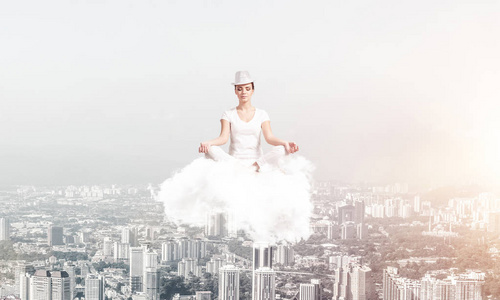 穿着白色衣服的女人闭上眼睛，集中注意力，在空中沉思云，背景上有城市景观的景色。