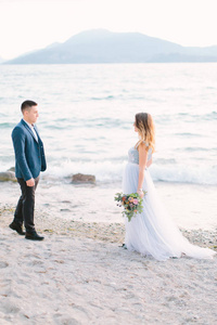年轻的新娘穿着漂亮的蓝色婚纱，在加德纳湖附近的海滩上走向新郎。 结婚日和爱情概念