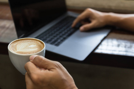 关闭男子手拿咖啡杯和打字笔记本电脑。 商人用笔记本电脑在咖啡店工作。 商业技术生活方式的概念。