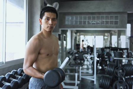 年轻的亚洲男人在健身房举起哑铃。 健康的生活方式和锻炼动机的概念。
