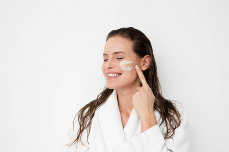 年轻的女人，完美的皮肤，在她的脸上涂上保湿霜。 妇女洗澡后的照片，白色浴袍和毛巾在白色背景。 护肤概念