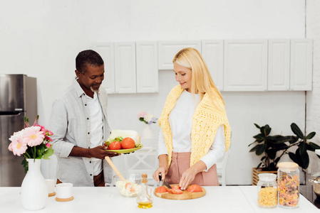成熟的女人在砧板上切西红柿，而非裔美国人男人拿着蔬菜盘子