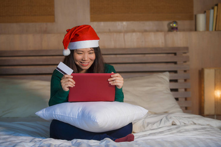 年轻快乐美丽的亚洲女性，戴圣诞帽，用信用卡和笔记本电脑上网购物，圣诞礼物和礼物，坐在床上，快乐兴奋