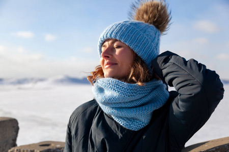 冬季背景上的肖像美女模特。美丽的现代年轻女子戴着蓝色针织帽温暖的双手，微笑着，看着镜头在冰封的海面上散步