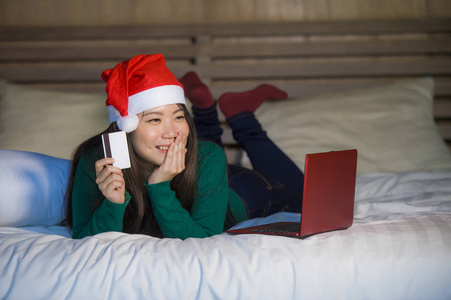 年轻快乐美丽的亚洲女孩戴着圣诞老人的圣诞帽，用信用卡和笔记本电脑进行网上购物，圣诞礼物和礼物坐在床上