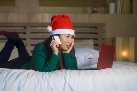 年轻快乐美丽的亚洲女孩戴着圣诞老人的圣诞帽，用信用卡和笔记本电脑进行网上购物，圣诞礼物和礼物坐在床上