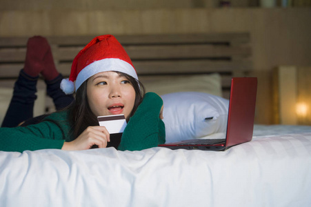 年轻快乐美丽的亚裔美国女孩戴圣诞帽，用信用卡和笔记本电脑上网购物，圣诞礼物和礼物，坐在床上快乐兴奋