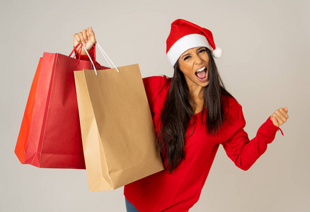 年轻的女人准备迎接圣诞节，带着红色的纸袋和圣诞老人的帽子，在准备圣诞购物礼物庆祝消费主义和销售理念时，隔离了灰色的背景。