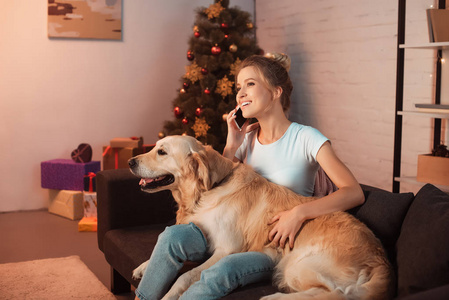 圣诞节那天，美丽的金发女郎和一只金毛猎犬坐在沙发上，在智能手机上聊天