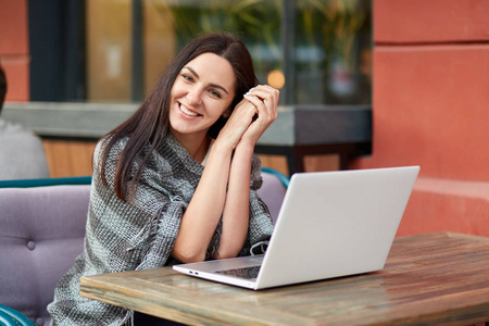 快乐的女性自由职业者，对户外餐厅的远程工作感到满意，肩膀上有被子，坐在打开的笔记本电脑前面，独自一人看起来很开心。 休闲与科技