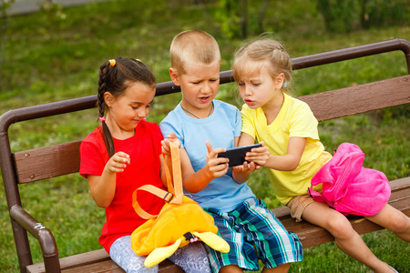 三个快乐的孩子在公园的长凳上玩智能手机