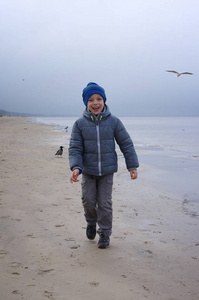 年轻的微笑男孩沿着冬天的海滩跑。快乐的孩子穿着温暖的灰色夹克。许多海鸥飞来飞去