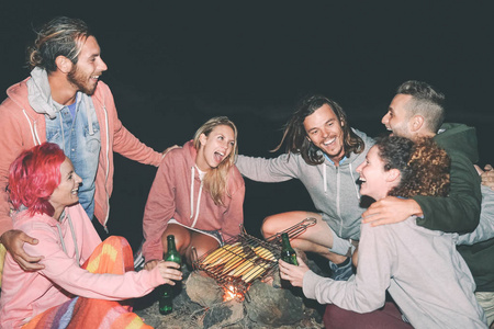 快乐的朋友在烧烤上煮新鲜的玉米蔬菜，户外的年轻人在露营的时候玩，晚上笑，喝啤酒，下一个篝火，年轻人的生活方式概念