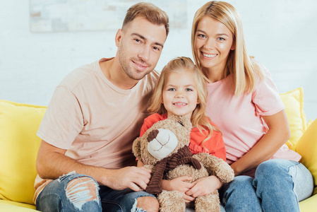 微笑的父母坐在沙发上看着相机，女儿抱着泰迪熊