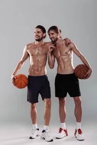 两个快乐的肌肉发达的赤膊的孪生兄弟站在灰色背景上，手持篮球