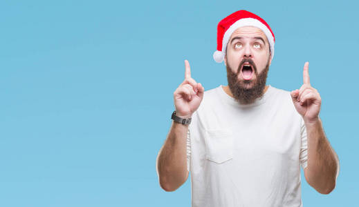 年轻的白种人嬉皮士戴着圣诞帽在孤立的背景上惊讶和惊讶地抬头，用手指和举起的手臂指着。
