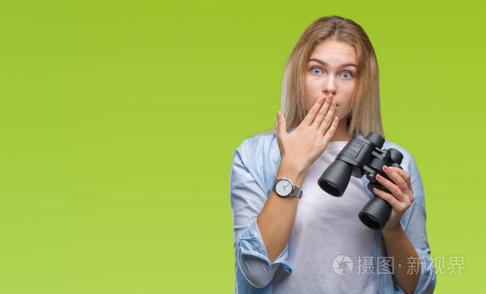 年轻的白种人女人拿着双筒望远镜在孤立的背景罩口上用手震惊,羞愧