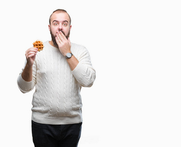 年轻的白种人时髦的男人在孤立的背景盖嘴上吃甜华夫饼，手因错误地表达恐惧而震惊，在沉默中害怕秘密概念