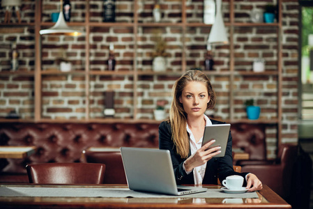 女商人坐在自助餐厅时使用平板电脑。 在桌子上，笔记本电脑和咖啡。
