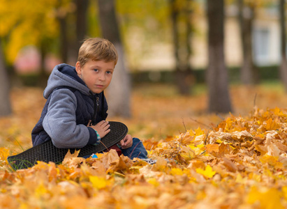 一个年轻孤独的男孩，他的滑板坐在公园里棕色的落叶里。
