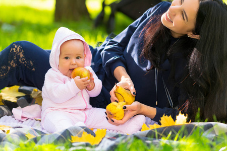 在秋天的公园里，她坐在草地上的地毯上，抱着一个成熟的金苹果，快乐的年轻妈妈嘲笑她的小女儿