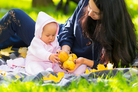年轻的母亲带着她的小女儿去看新鲜的秋苹果，因为他们在公园的近景草地上的地毯上一起放松
