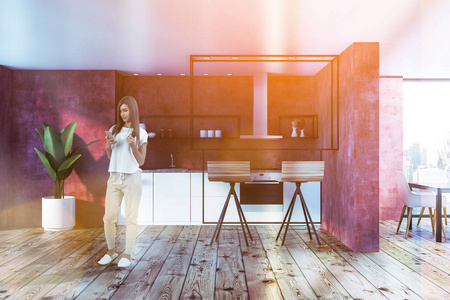 现代厨房里的女人，红色的墙壁，木制的地板，白色和灰色的台面，烤箱和水槽，两个凳子站在酒吧桌子附近。 调色图像
