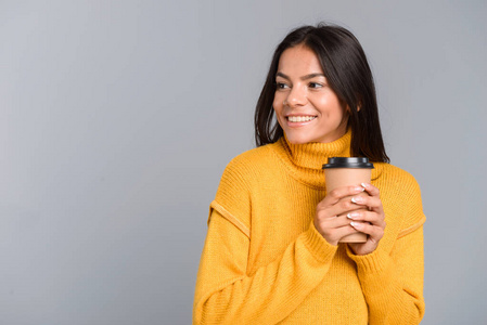 一位微笑的年轻女子的肖像，她穿着毛衣，手里拿着外卖的咖啡杯，在灰色的背景下孤立地看着远方