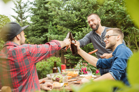 快乐的朋友坐在花园的桌子旁，享受食物和啤酒，品尝啤酒瓶。