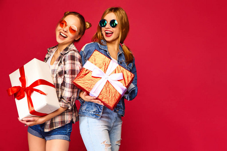 圣诞节的概念。两个微笑的漂亮女人穿着时尚的衣服。女孩在红色的背景上摆姿势。带大礼盒的模特。准备庆祝。最好的朋友的节日