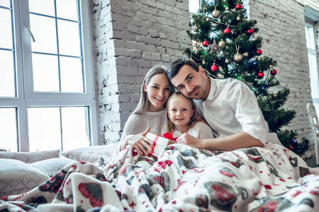 一家人坐在圣诞树附近的床上。妈妈，爸爸和宝宝在卧室里玩得很开心。人们在家放松。寒假圣诞节及新年概念