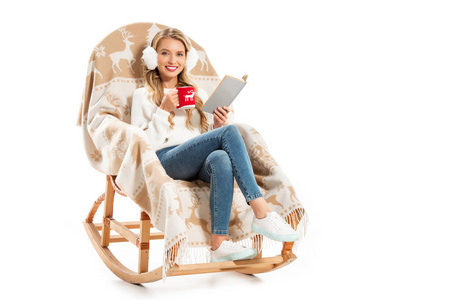 年轻的微笑的女人拿着书和咖啡杯，坐在摇椅上，与白色隔离