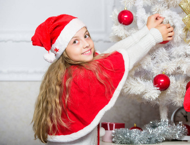 家庭传统概念。用红色的球饰品装饰圣诞树的儿童。装饰圣诞树的女孩孩子。珍爱人的假期活动。孩子在圣诞老人帽子装饰圣诞树