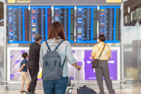 亚洲女性旅客的后侧，行李站在飞行板上，在现代机场旅行和运输概念的飞行信息屏幕上办理登机手续
