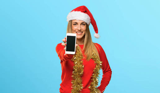 一位身着圣诞服装的金发女人看着相机微笑着使用蓝色背景的手机
