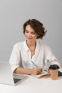 年轻快乐的商业女性的形象，在灰色的墙壁背景上，坐在桌子上，使用笔记本电脑。