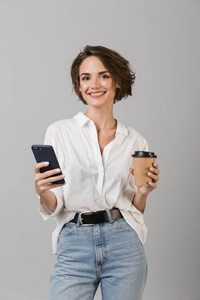 快乐的年轻商务女性的形象，在灰色的墙壁背景下，通过手机喝咖啡聊天。