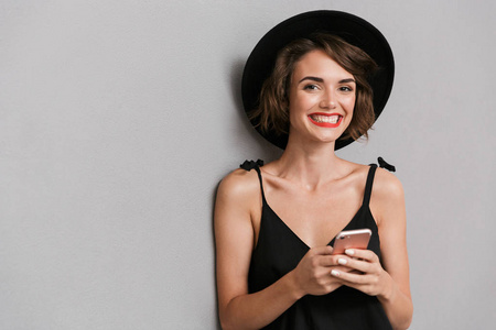 照片中，20岁的快乐女人穿着黑色连衣裙和帽子，微笑着看着相机，拿着智能手机，被隔离在灰色背景上