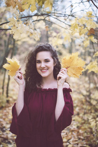 一个美丽的女孩，一头卷曲的黑发，在秋天公园的栗色顶部，微笑着看着相机，拿着枫叶