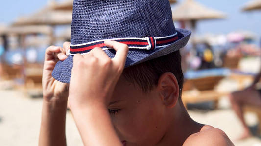 可爱而英俊的男孩带着帽子在海滩上摆姿势，微笑着，选择性地集中注意力