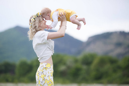 一位快乐的母亲抱着她刚出生的儿子站在高地的一片盛开的洋甘菊田野里。 一家人的母亲和婴儿在群山和蓝天的背景下，在一片盛开的雏菊的白