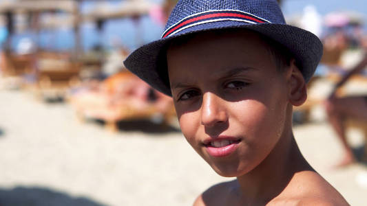 可爱而英俊的男孩带着帽子在海滩上摆姿势，微笑着，选择性地集中注意力