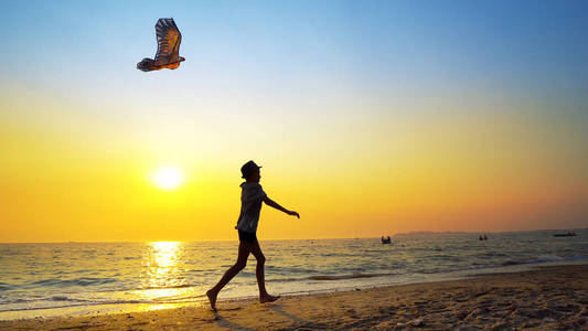 一个带帽子的小男孩的剪影一个空旷的海滩上放风筝，被夏日日落的阳光拍摄