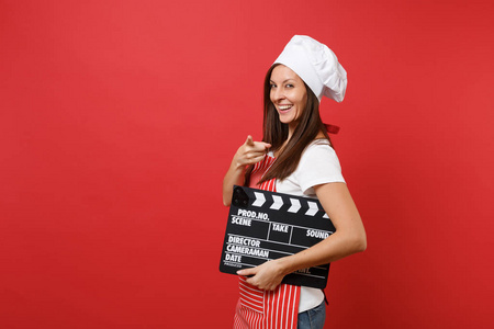 家庭主妇女厨师或面包师穿条纹围裙，白色T恤，烤厨师帽子隔离在红墙背景上。 女人拿着经典的黑色电影制作剪贴板。 模拟复制空间概念