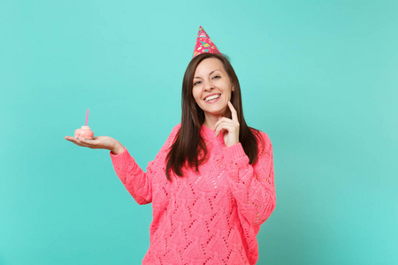 微笑的年轻女子，穿着针织粉色毛衣，生日帽，把手手指撑在下巴上，手拿蛋糕，蓝墙背景上的蜡烛。 人们的生活方式概念。 模拟复制空间