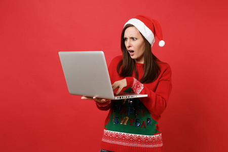令人震惊的年轻圣诞老人女孩戴着圣诞帽，在笔记本电脑上打字，电脑被隔离在明亮的红色背景上。 新年快乐，2019年庆祝节日聚会的理念