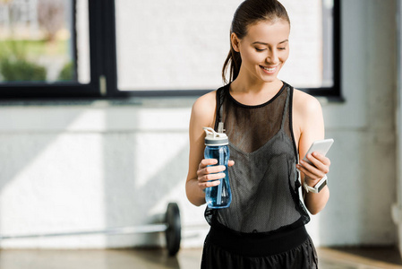 微笑的女运动员拿着带水的运动瓶，在健身房里用智能手机