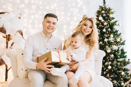 圣诞树和白色棉花背景上美丽的年轻家庭的肖像。 有魅力的父母和小儿子新年礼物