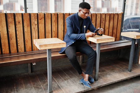 令人惊讶的是，非裔美国人穿着蓝色夹克，胸针黑色高领毛衣和眼镜摆在街上。 时髦的黑人拿着手机坐在桌子后面。