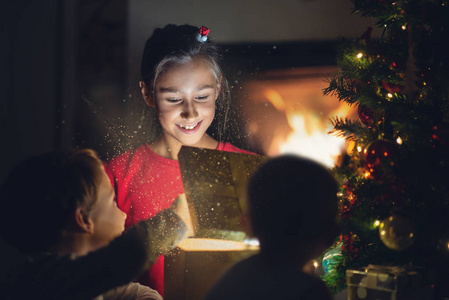 小女孩高兴地微笑着，她打开圣诞树下的金色礼盒，她的兄弟姐妹在壁炉的背景下点火。